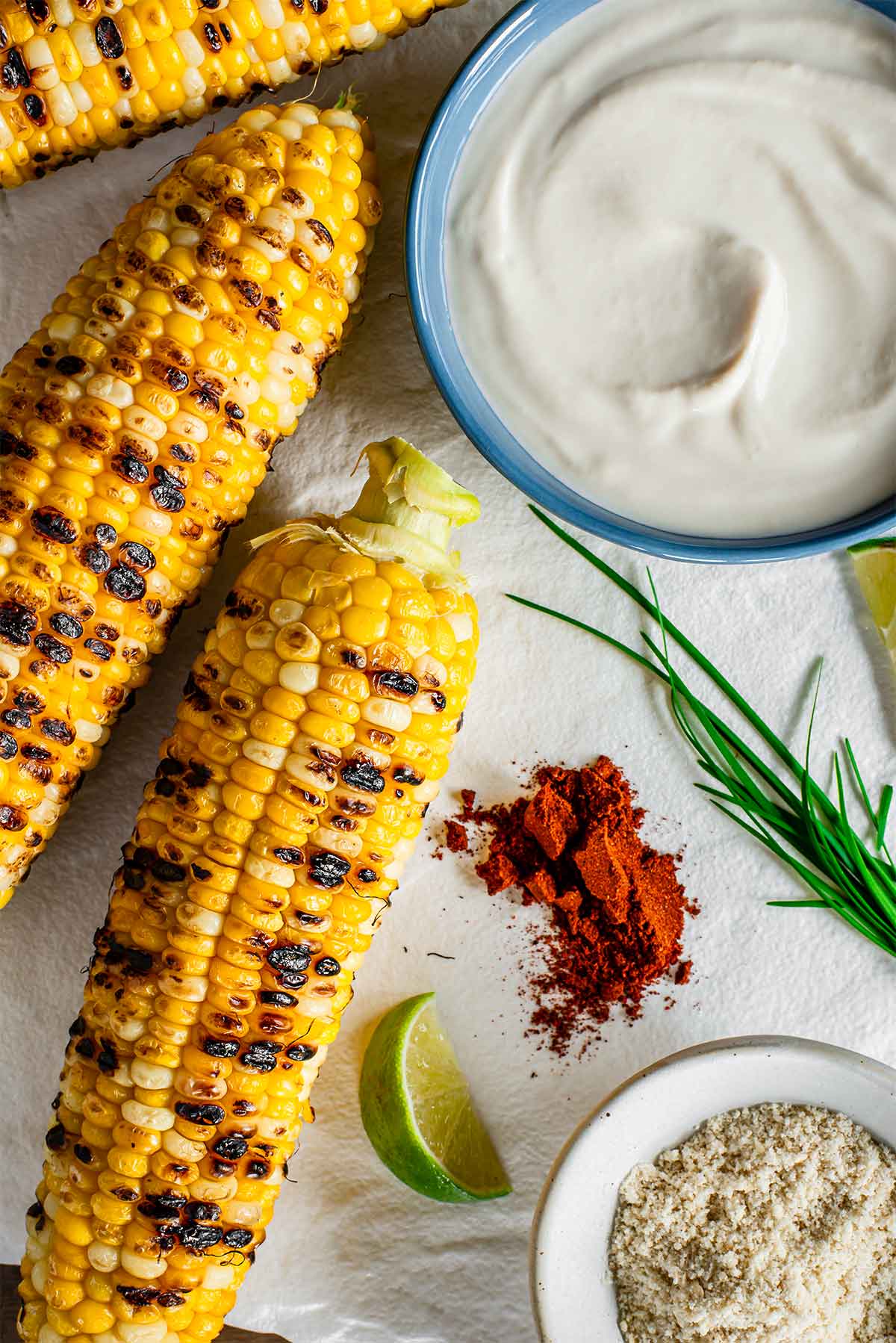 Top down view of grilled corn on the cob resting beside a bowl of thick tofu based mayonnaise, a small dish of cashew based "cheese", ancho chili powder, lime wedges, and garlic chives.