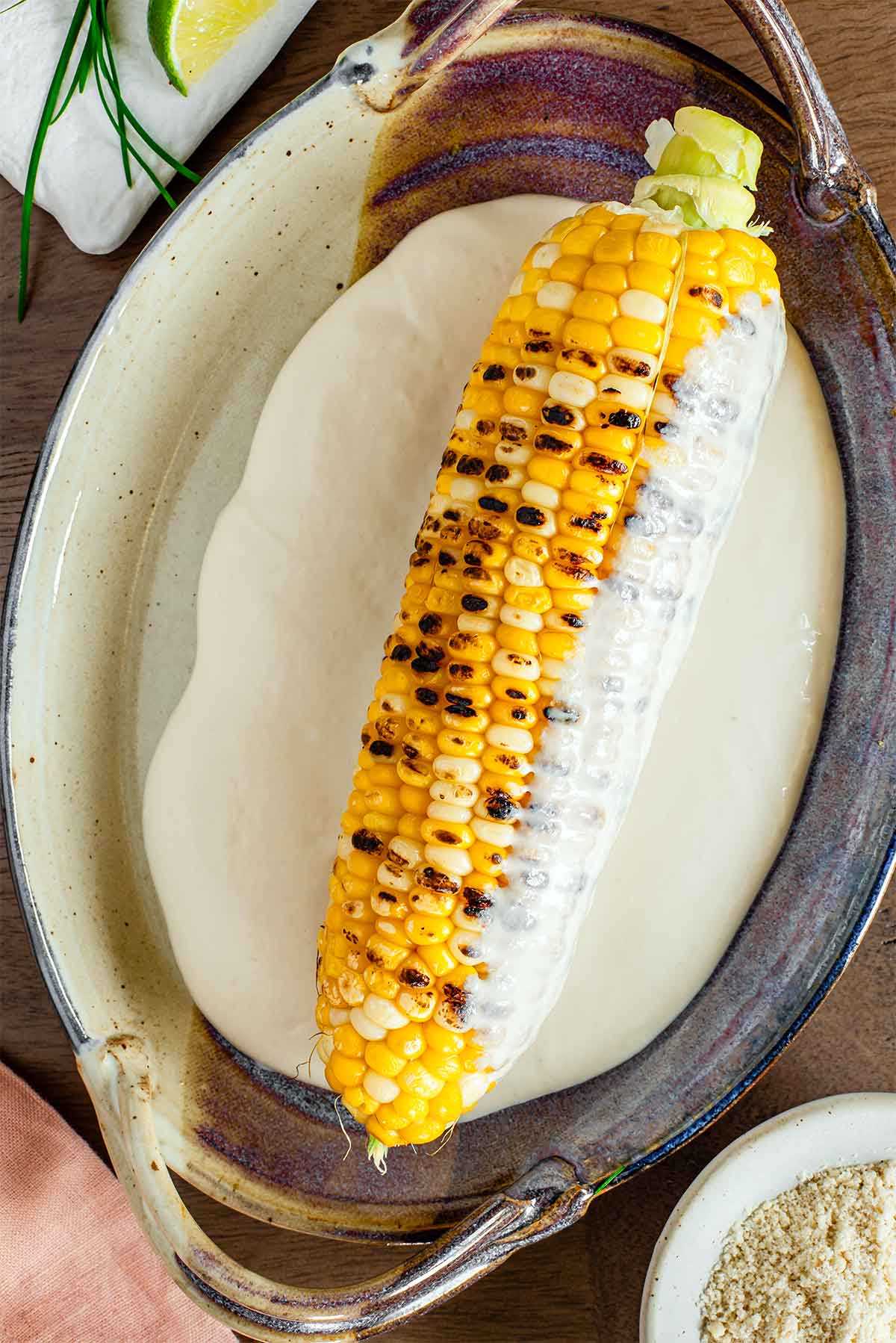 Top down view of a grilled cob of corn resting in a shallow dish of creamy tofu based mayonnaise. The cob has two inches of stem still attached, nicely charred grill lines, and has been half rolled in the creamy sauce.