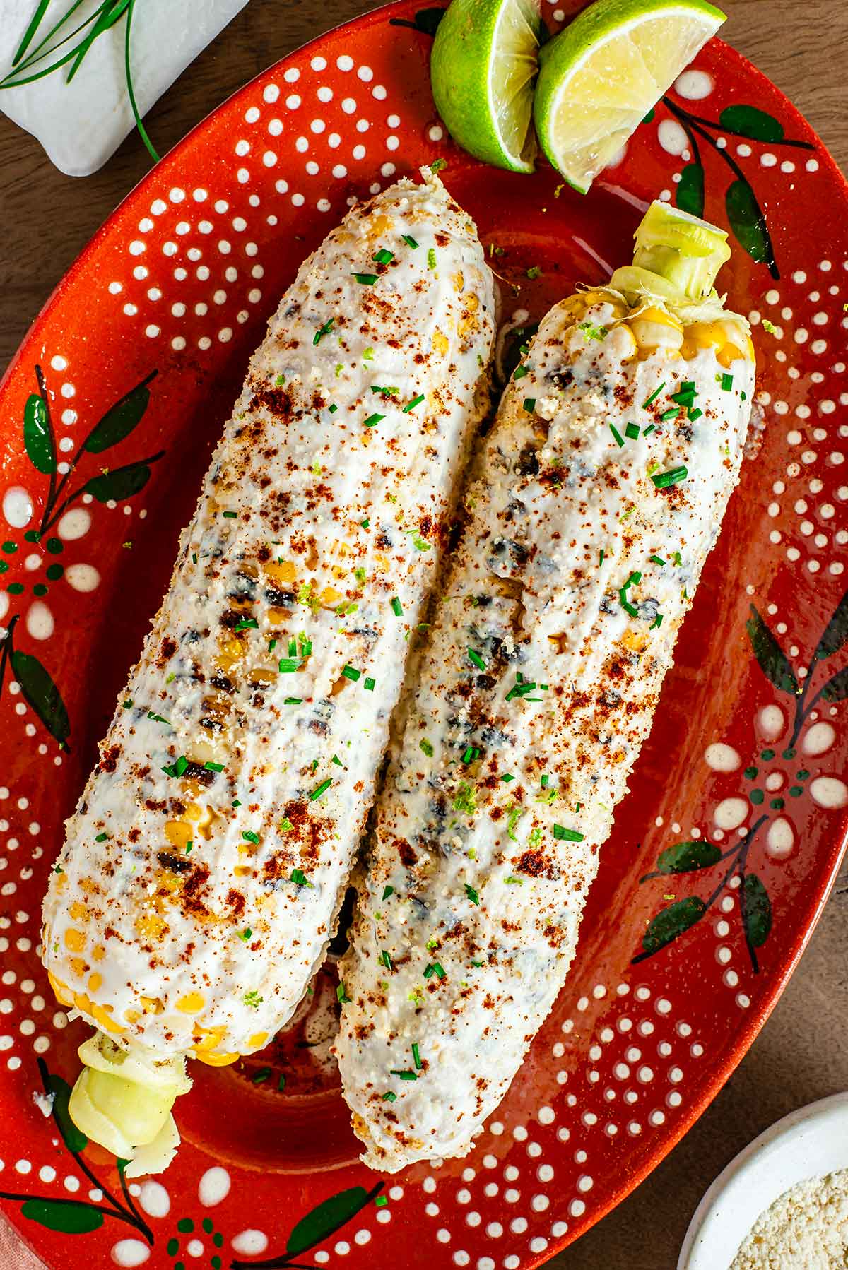Top down view of two cobs of vegan Mexican street corn on a decorative clay platter. The grilled cobs of corn are coated in creamy and cheesy toppings, garnished with chives and chili powder, and served alongside lime wedges.