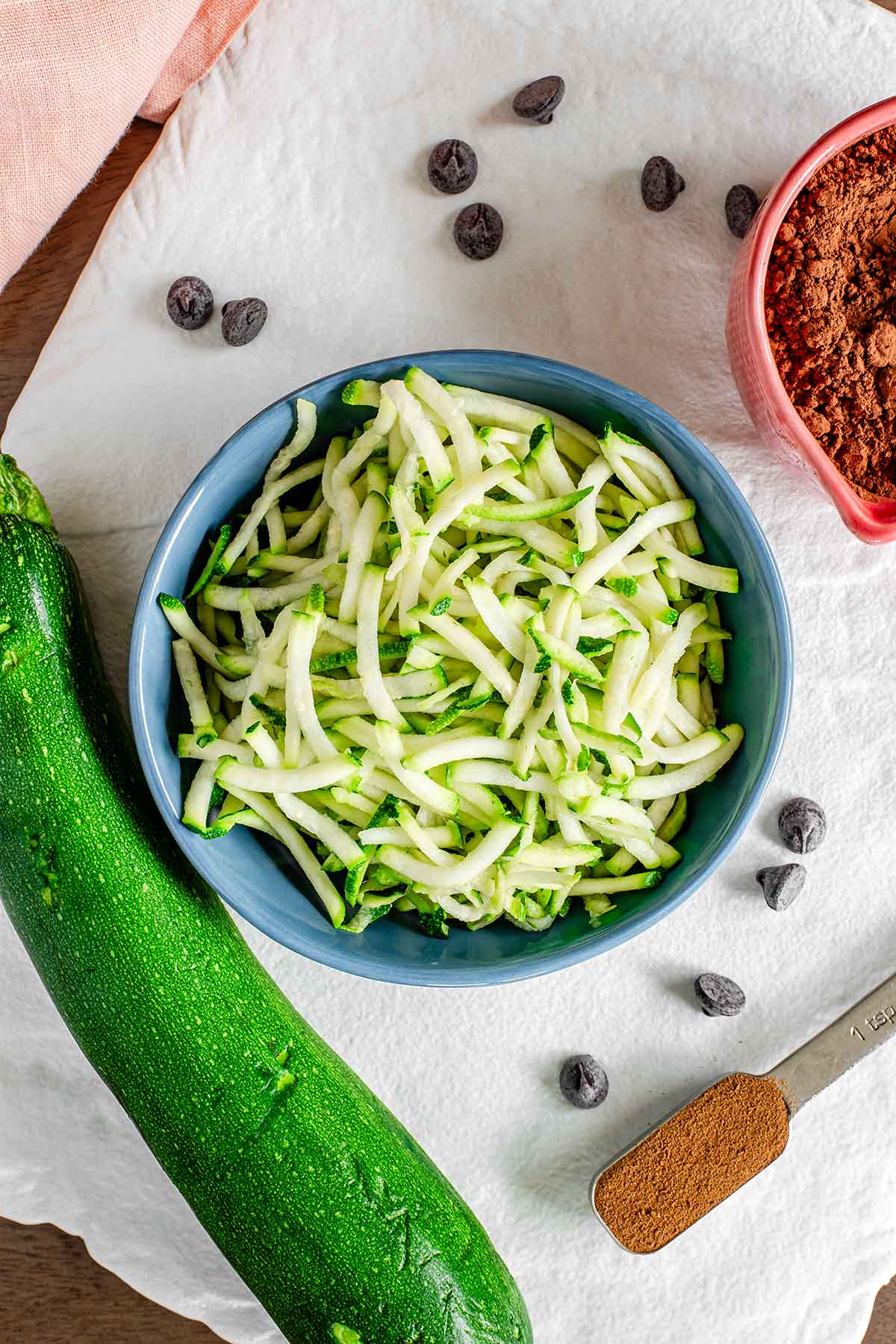 Top down view of shredded zucchini in a small bowl on a white tray. An intact green zucchini rests beside the bowl. Cocoa powder, chocolate chips, and espresso powder surround the zucchini.