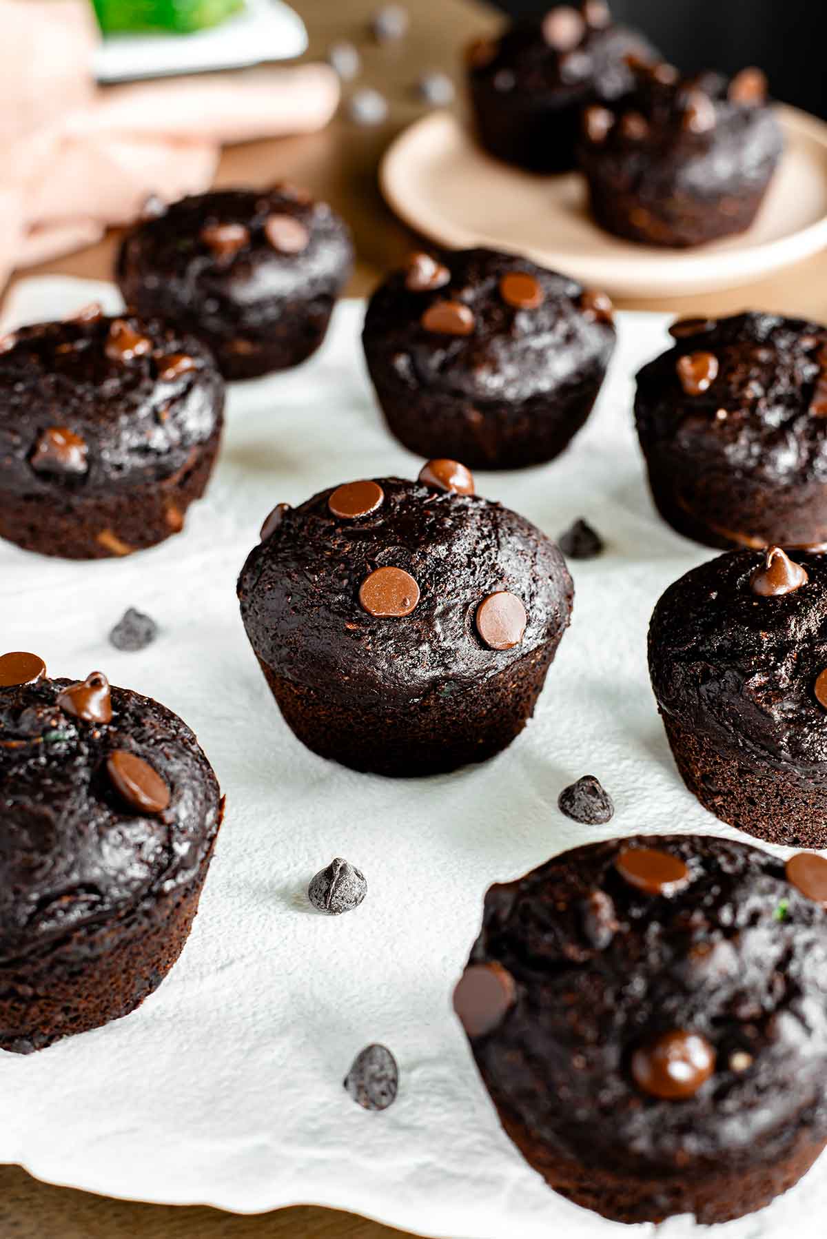 Side view of a chocolate zucchini muffins on a white tray. The muffins are smaller homemade style muffins that are dark chocolate brown and speckled with chocolate chips. A green zucchini can be seen in the background.