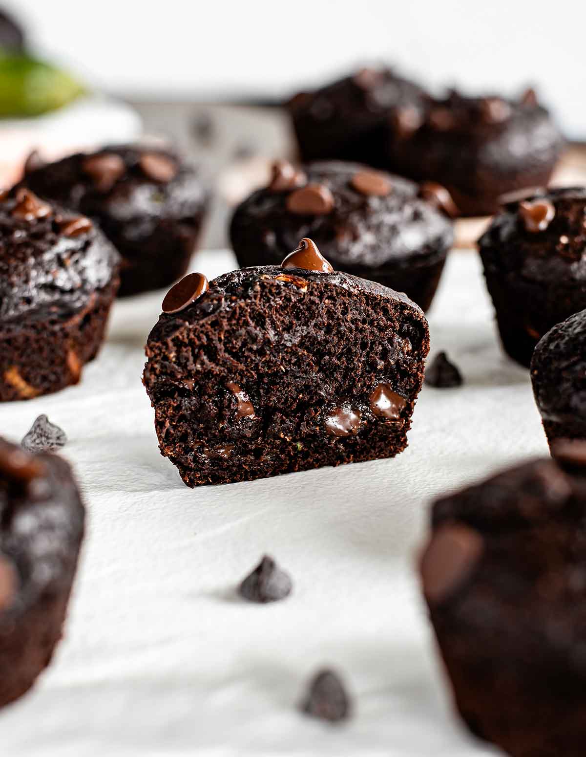 Half of a chocolate zucchini muffin rests on a white tray. The cut muffin shows that the crumb is tender and moist, the chocolate chips speckled throughout are gooey, and the zucchini is practically invisible in the dark chocolate cake. 