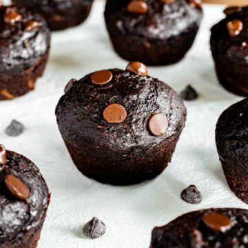Side view of a chocolate zucchini muffins on a white tray. The muffins are smaller homemade style muffins that are dark chocolate brown and speckled with chocolate chips.