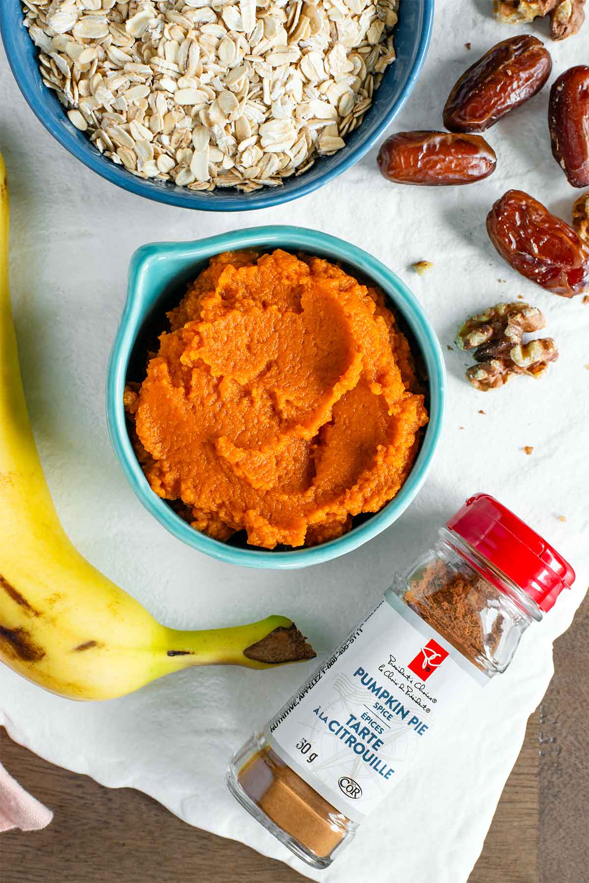 Top down view of ingredients on a white tray. Pumpkin purée fills a measuring cup in the centre of the tray, rolled oats, a ripe banana, dates and walnuts surround. A small jar of pumpkin pie spice also lays on the tray.