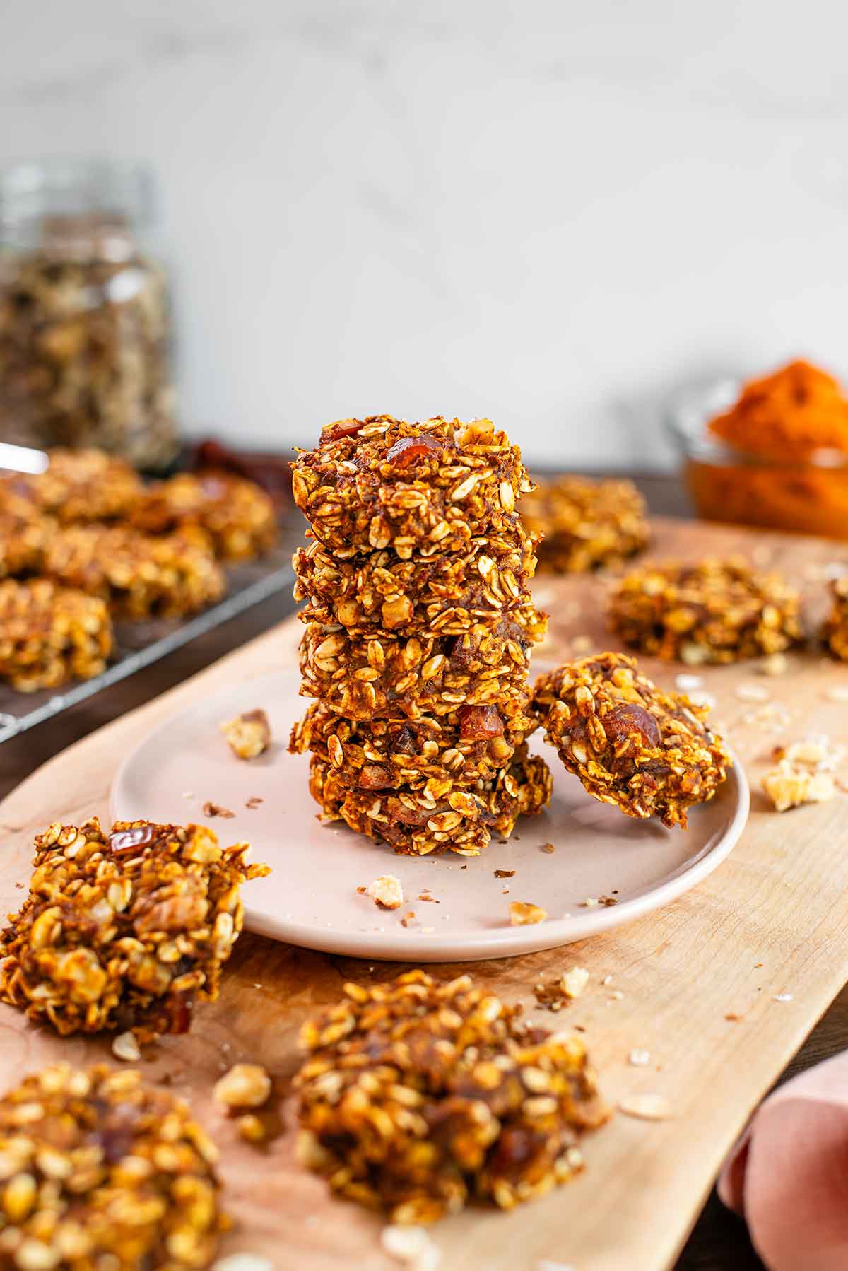 Side view of a stack of healthy pumpkin cookies. More cookies are scattered on a wooden tray. The cookies are densely packed with oats, dates and walnuts.