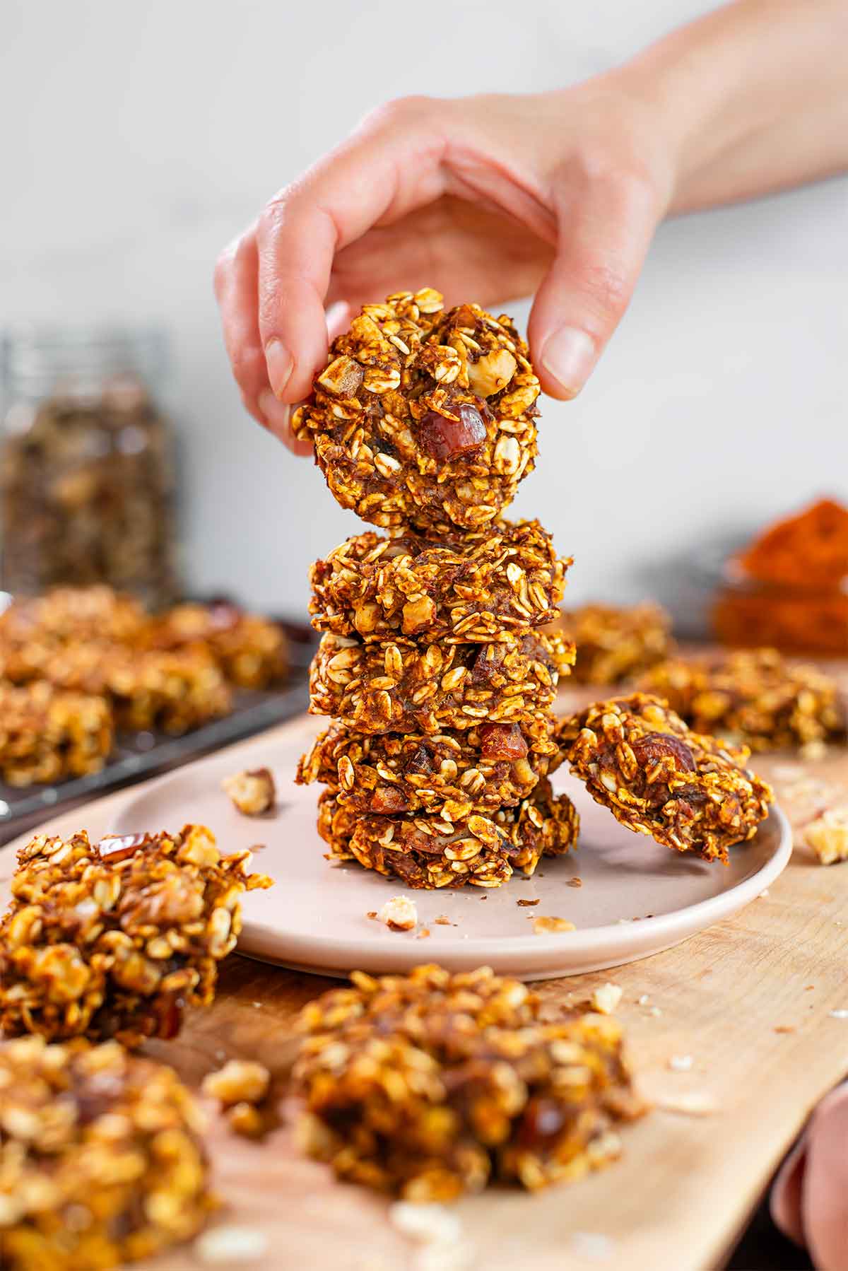Side view of a hand lifting a healthy pumpkin cookie from a tall stack. The oat cookies are orange from the colour of the pumpkin and speckled with chopped dates and walnuts. More cookies are scattered around a wooden tray.