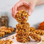 Side view of a hand lifting a healthy pumpkin cookie from a tall stack. The oat cookies are orange from the colour of the pumpkin and speckled with chopped dates and walnuts.