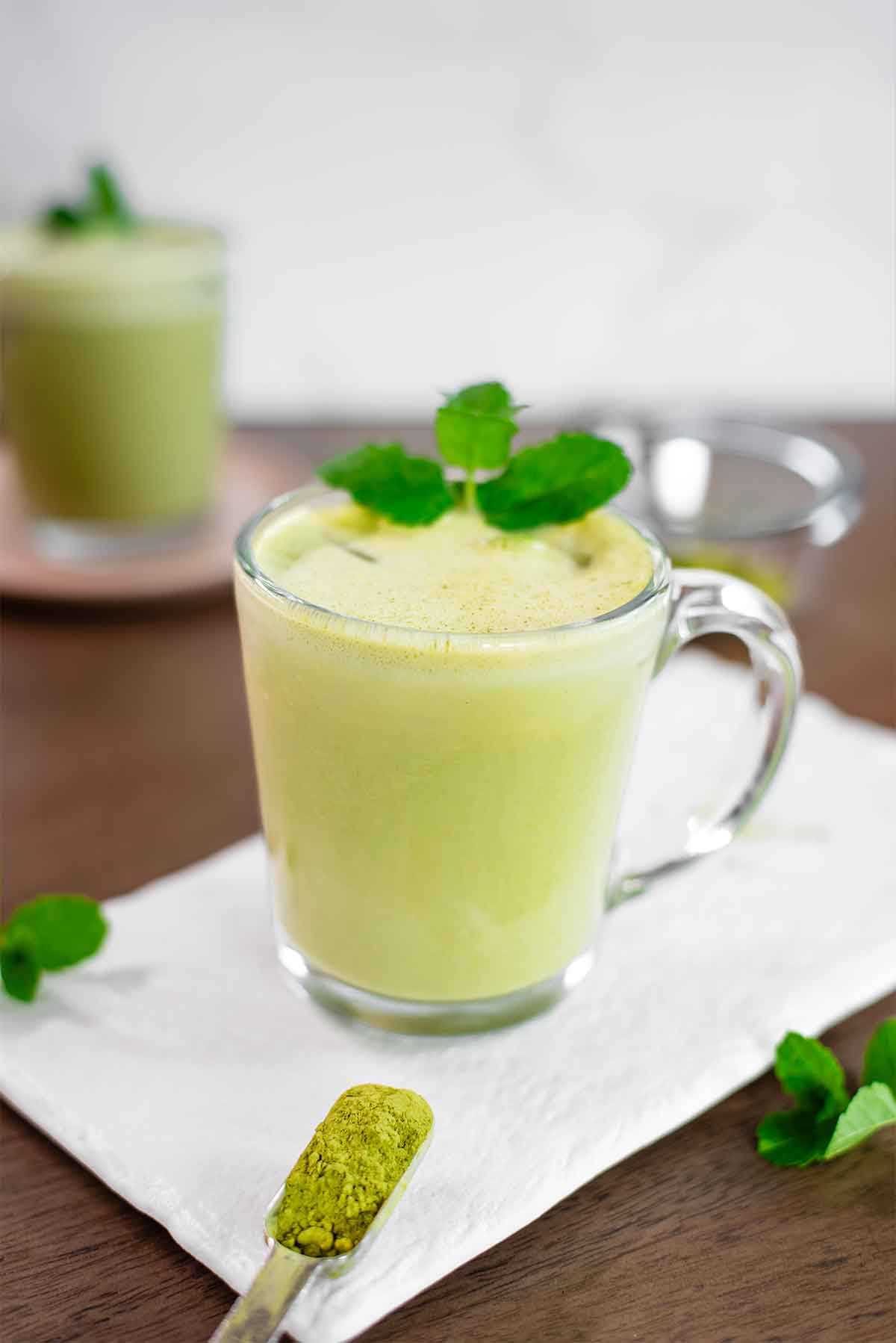 Side view of a creamy iced mint matcha latte in a clear mug. The latte is a pale green colour with foam on top, ice cubes visible in the mug, and is garnished with fresh mint leaves. A teaspoon of matcha powder rests in front of the mug on a white tray. 