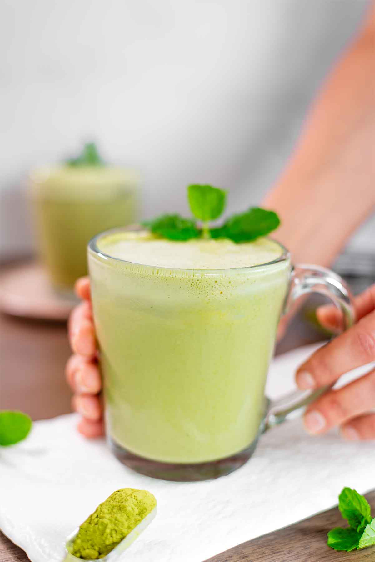Side view of hands placing a mint matcha latte on a white tray. The latte is garnished with fresh mint leaves.