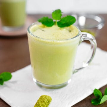 Side view of a creamy iced mint matcha latte in a clear mug. The latte is a pale green colour with foam on top, ice cubes visible in the mug, and is garnished with fresh mint leaves.