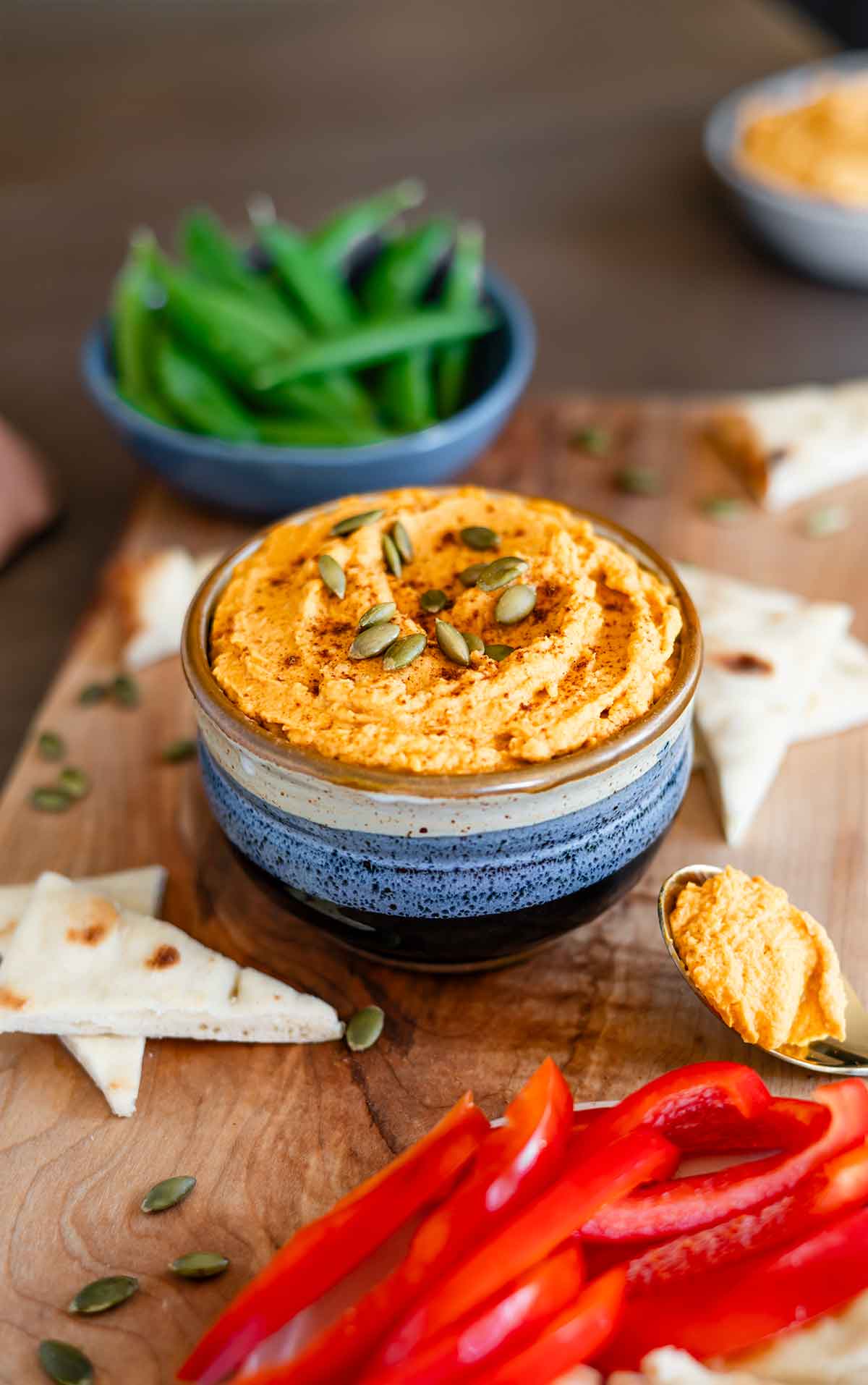 Side view of pumpkin hummus in a small bowl surrounded by cut veggies and toasted flatbread.