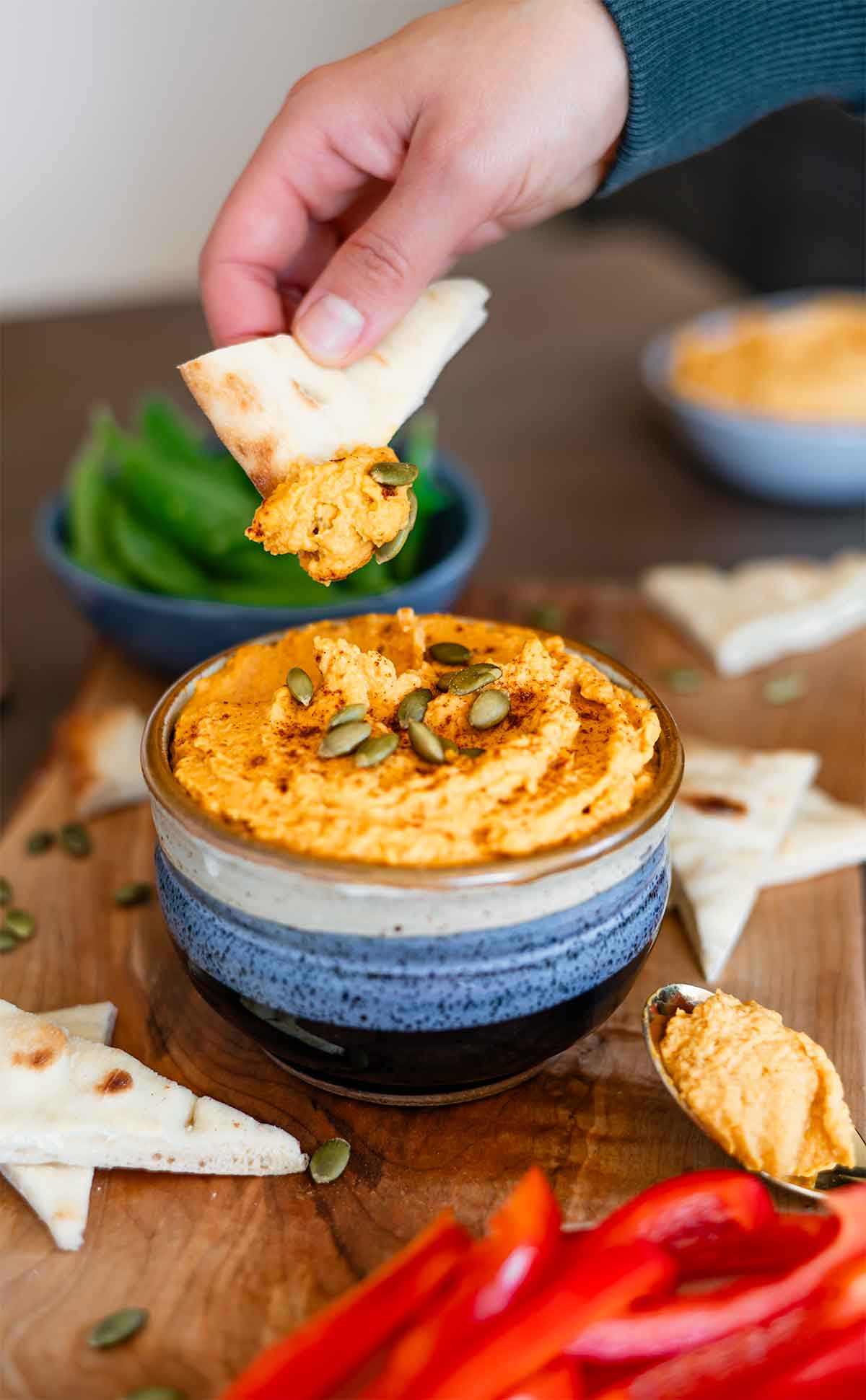 Side view of a hand dipping a piece of flatbread into a creamy orange coloured hummus topped with pumpkin seeds.