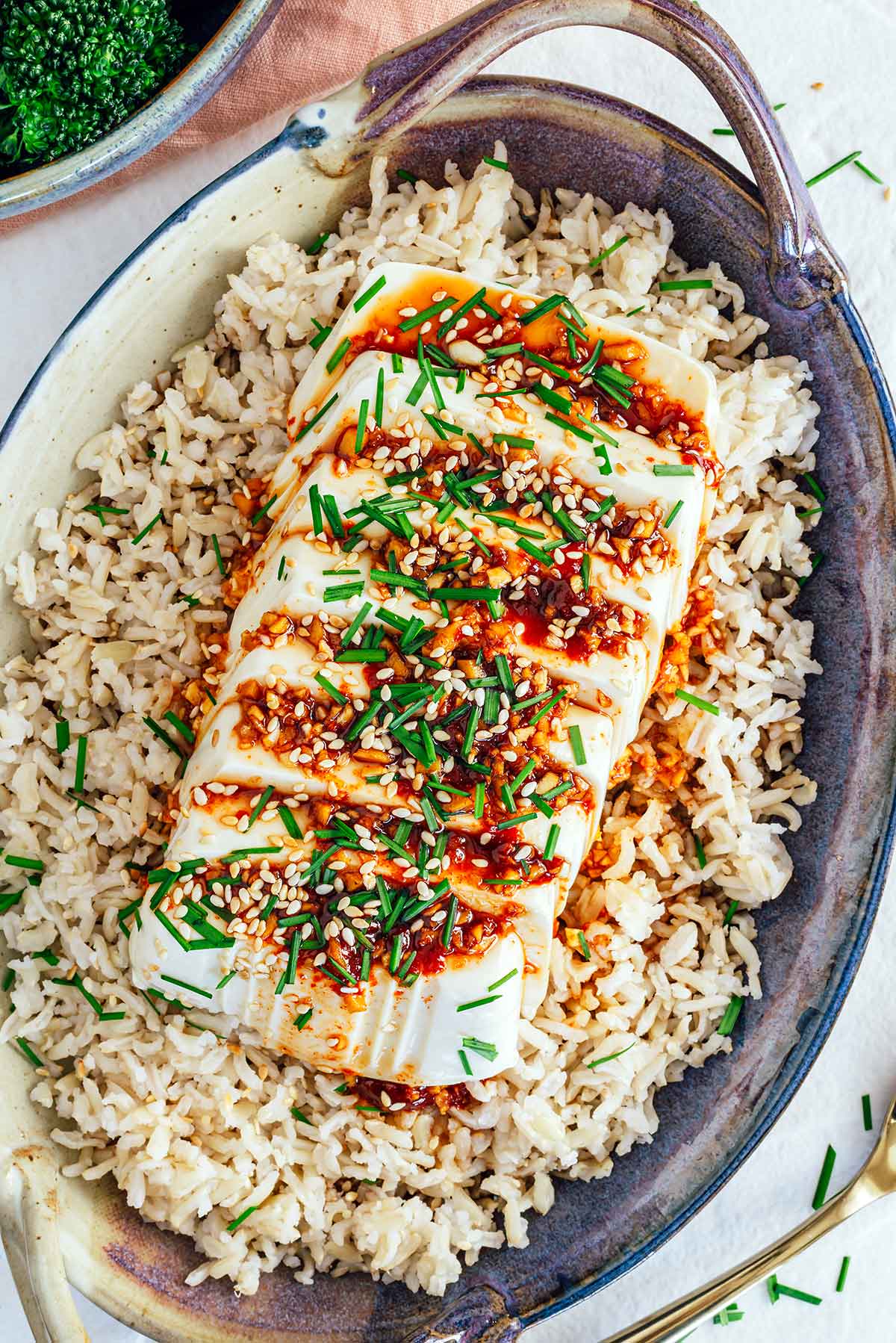 Top down view of cold soft tofu atop a bed of rice. The tofu is sliced widthwise into 8 slices and a red sauce is drizzled over top. The tofu is garnished with toasted sesame seeds and garlic chives.