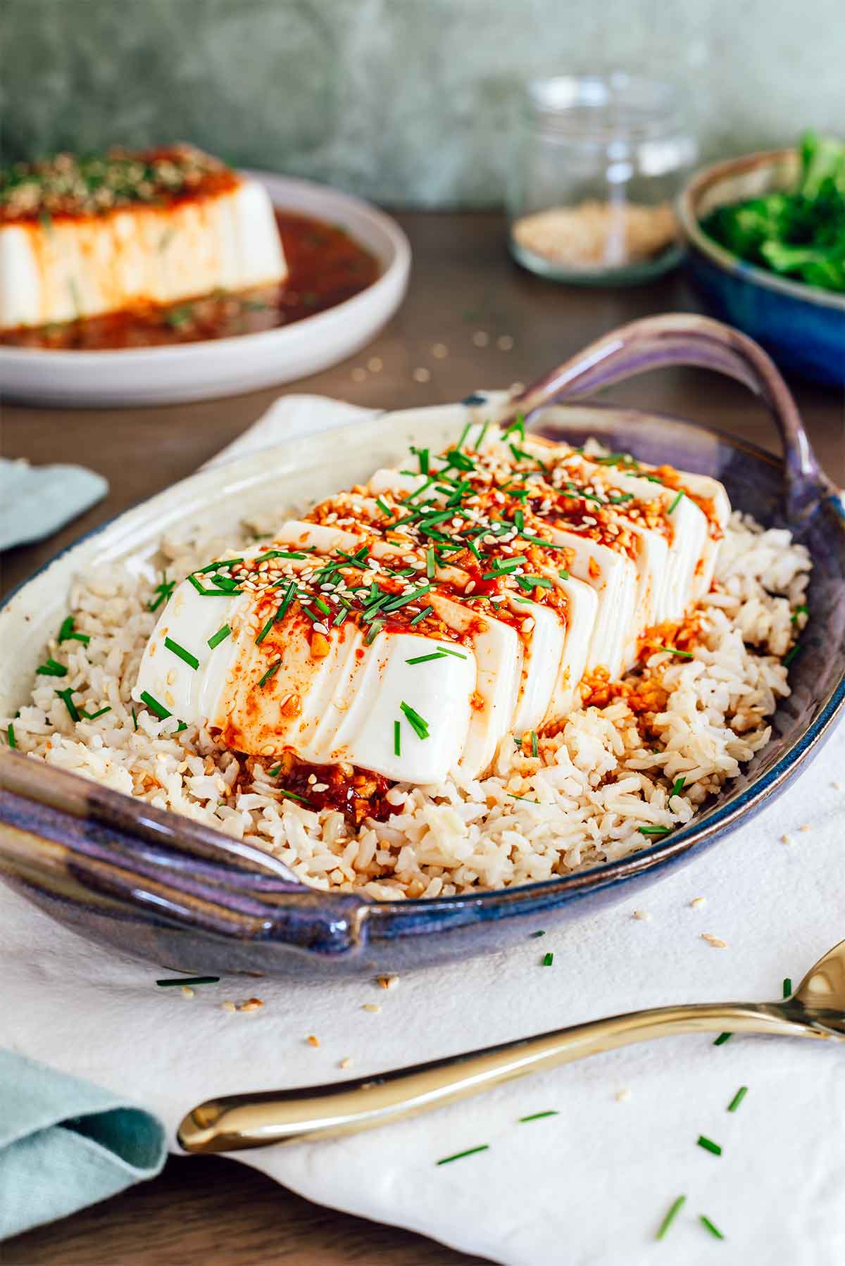 Side view of cold soft tofu sliced and sitting atop a bed of rice. The tofu is coated in a red sauce and garnished with toasted sesame seeds and garlic chives.