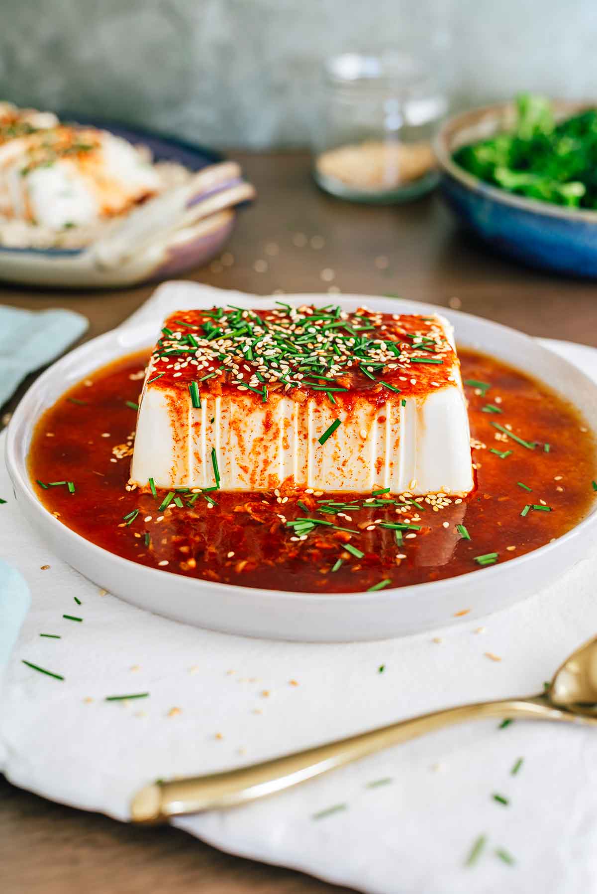A plate rests on a white tray holding a brick of cold soft tofu covered in a red sauce. The soy sauce and gochujang sweet and spicy dressing runs over the side of the tofu. Toasted sesame seeds and chopped garlic chives garnish the top of the tofu.