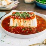 Side view of a brick of cold soft tofu covered in a red sauce. The soy sauce and gochujang sweet and spicy dressing runs over the side of the tofu. Toasted sesame seeds and chopped garlic chives garnish the top of the tofu.