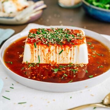 Side view of a brick of cold soft tofu covered in a red sauce. The soy sauce and gochujang sweet and spicy dressing runs over the side of the tofu. Toasted sesame seeds and chopped garlic chives garnish the top of the tofu.