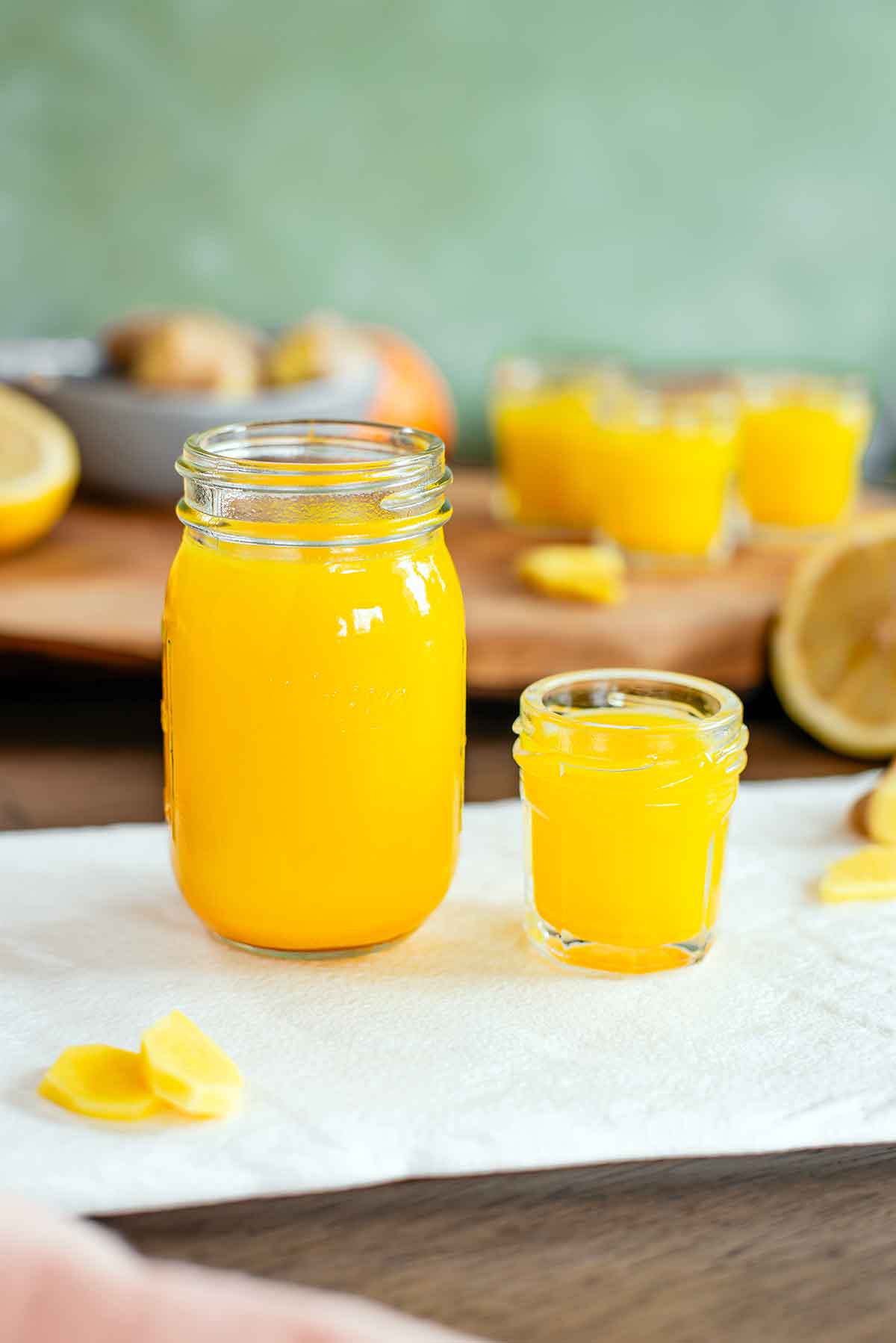 Side view of a larger jar of ginger citrus juice beside a smaller 1 oz shot glass of the liquid. The liquid is bright yellow and more shots and ingredients can be seen in the background.