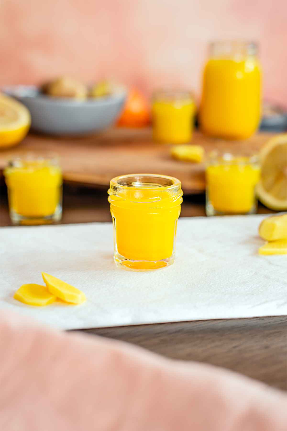 Side view of a ginger shot in a small 1 oz glass jar. The ginger shot is bright yellow and sliced ginger root lays beside it. More ginger shots and ingredients fill the background of the image.