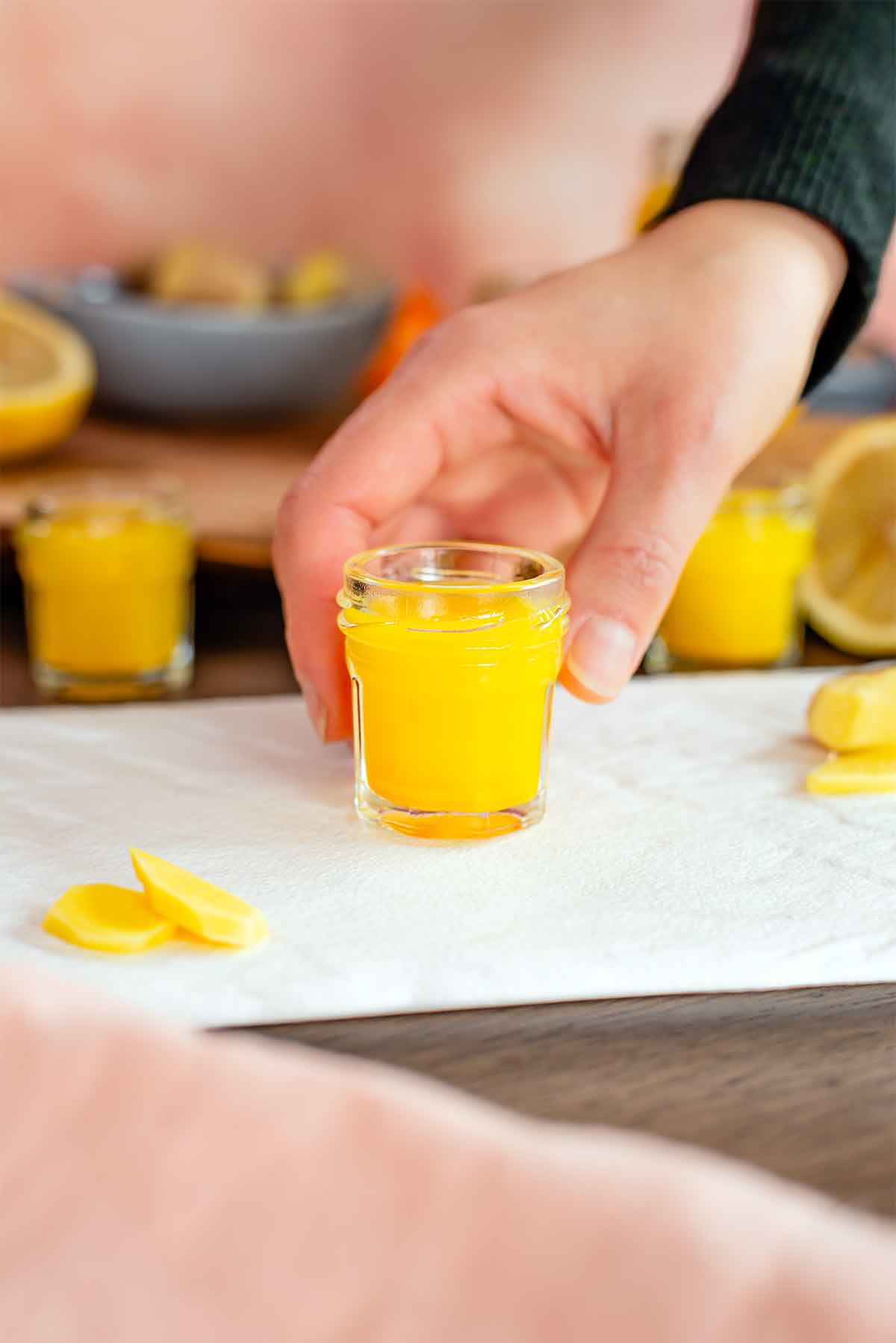 Side view of a hand reaching for a small ginger shot in glass jar. More ginger shots and ingredients fill the background of the image.