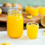 Side view of a larger jar of ginger citrus juice beside a smaller 1 oz shot glass of the liquid. The liquid is bright yellow.