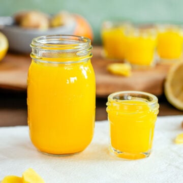 Side view of a larger jar of ginger citrus juice beside a smaller 1 oz shot glass of the liquid. The liquid is bright yellow.